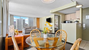 A modern living room with a glass dining table, wooden chairs, a sofa, and a view of high-rise buildings.