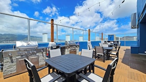 An outdoor deck with a barbecue area, tables, and chairs, overlooking a body of water and distant mountains.