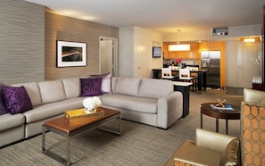 Ein modernes Hotelzimmer mit Ecksofa, Couchtisch, kleinen Beistelltisch und Essbereich, im Hintergrund eine Küche.
