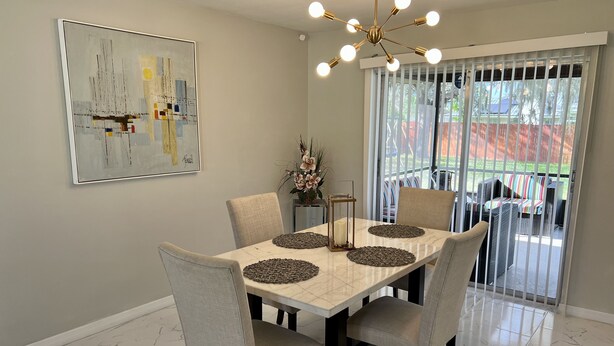 Un coin repas avec une table en marbre, des chaises beiges, un lustre moderne et une grande fenêtre équipée de stores.