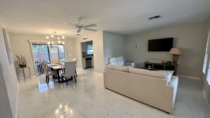Un vaste salon avec un grand canapé blanc, un coin repas avec une table et des chaises, une cuisine à l’arrière-plan et un ventilateur de plafond.