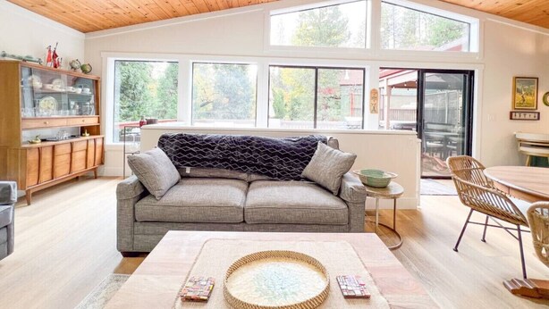 A cozy living room with a wooden ceiling, a grey sofa, a coffee table, and a dining area with wicker chairs.