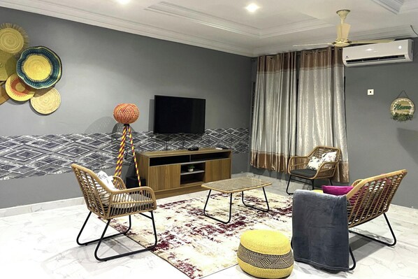 A modern living room with a flat-screen TV, two wicker chairs, a small ottoman, and a patterned rug.