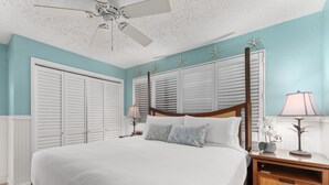 A bedroom with a large bed, a wooden nightstand, a ceiling fan, and a window with shutters. - Comfort of home, ground floor with in unit laundry (Kihei)