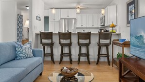 A modern kitchen with white cabinets, a stainless steel refrigerator, and a ceiling fan. There are four bar stools and a glass coffee table with a bowl of snacks. - Comfort of home, ground floor with in unit laundry (Kihei)