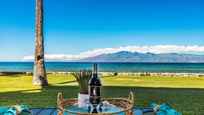 A table with a bottle of wine, glasses, and a plant, set against a scenic view of the ocean and mountains. - Direct oceanfront views, remodeled condo (Lahaina)
