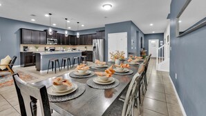 A modern kitchen with dark cabinets, a central island, and a dining area set for six with a long table and six chairs. - Windsor at Westside Resort 9-Bedroom Pool/Spa Home (Kissimmee)