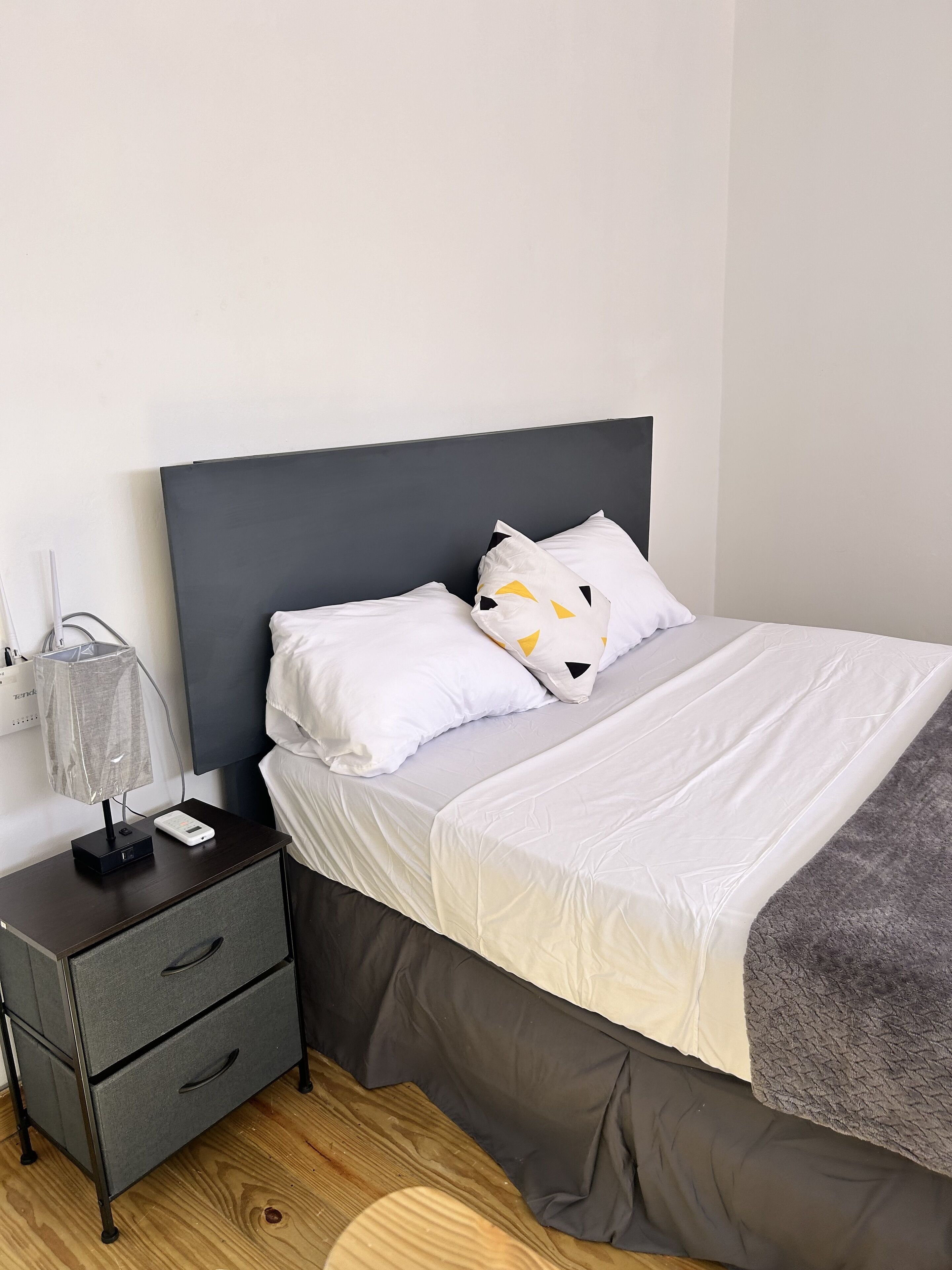 Una cama bien hecha con ropa blanca y una manta gris, una mesita de noche con lámpara y regleta, y un tocador gris con cajones.