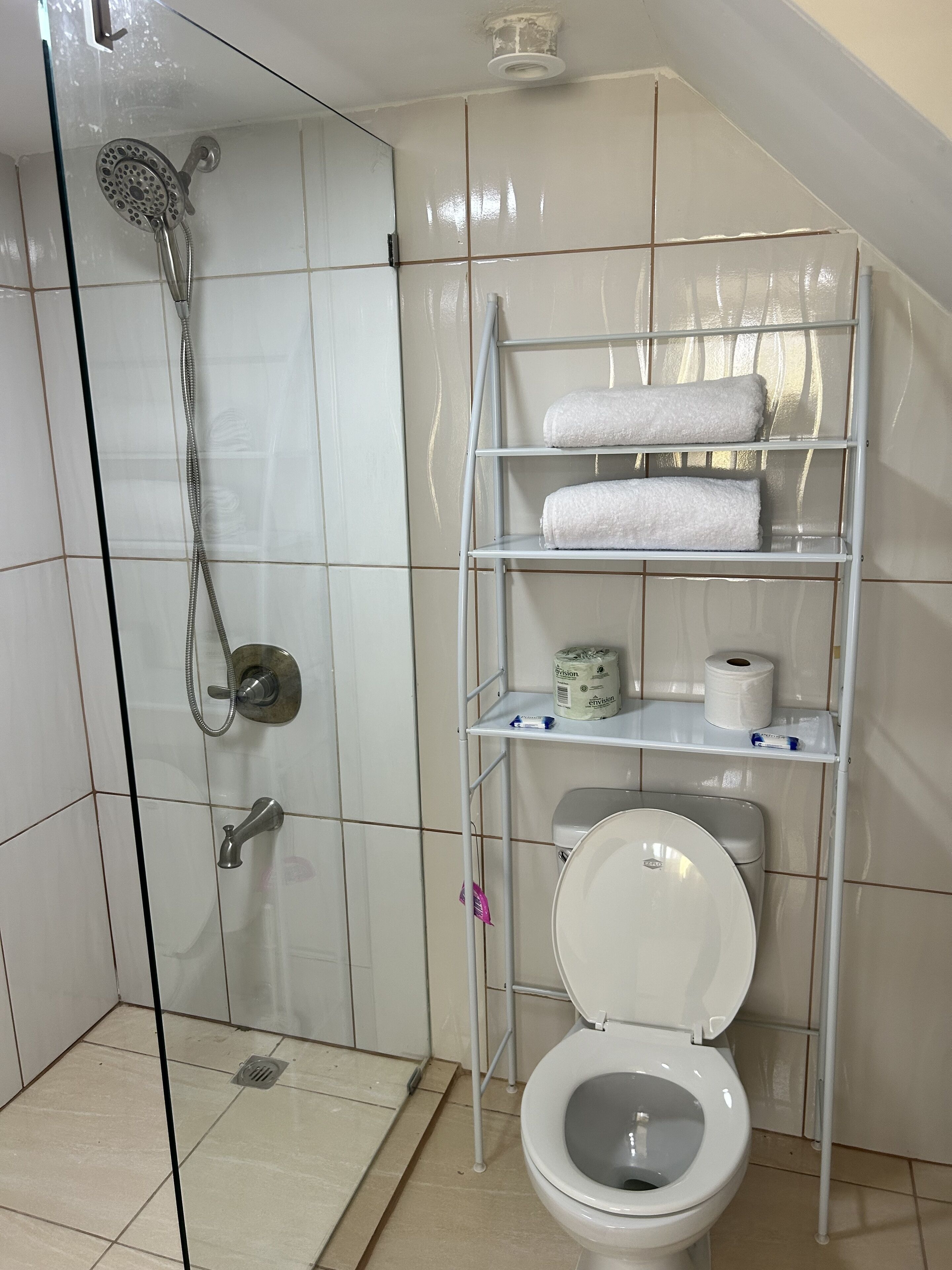 Casa de banho com chuveiro, sanita e prateleiras com toalhas e papel higiénico.