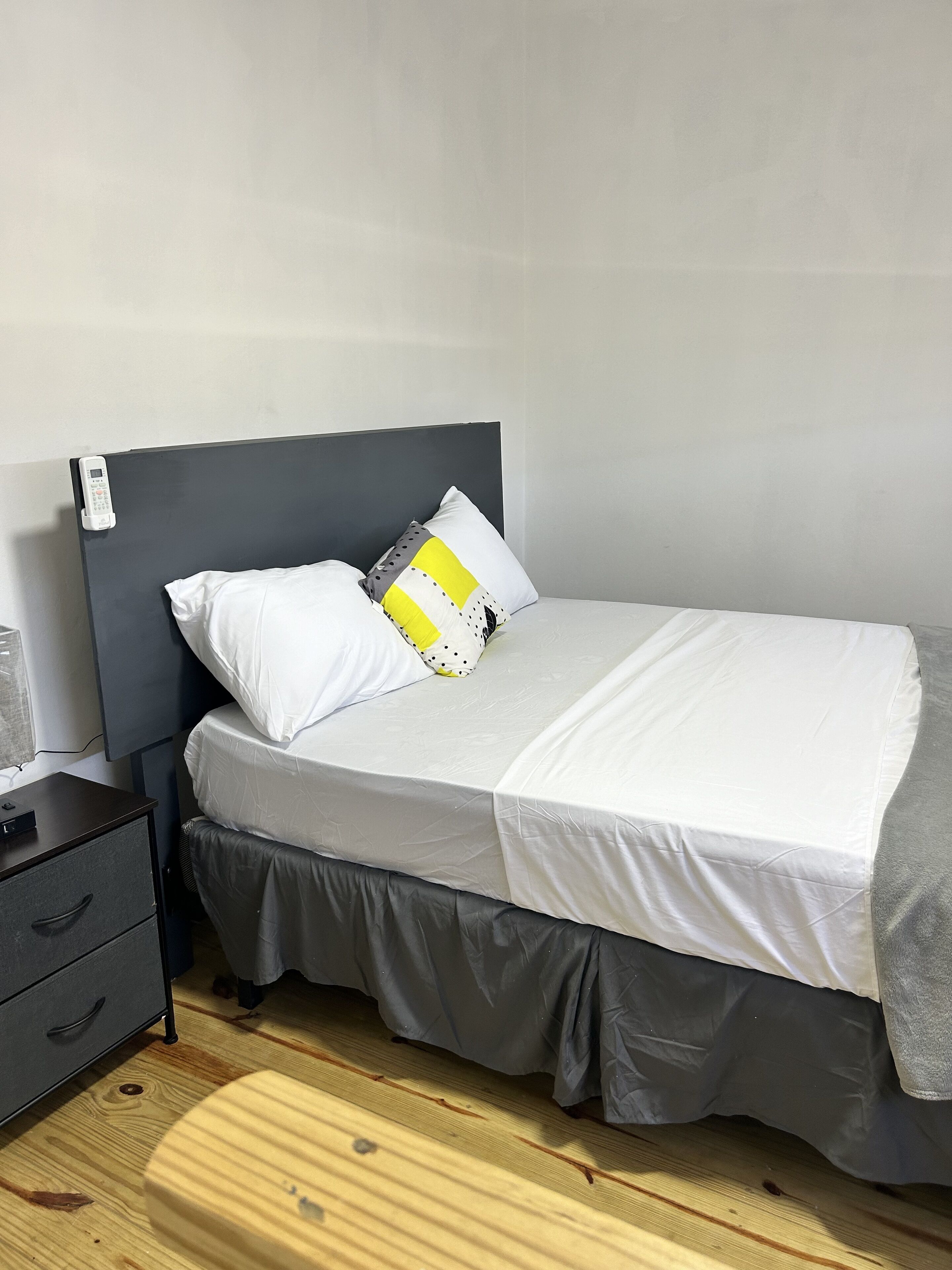 A neatly made bed with white bedding and a grey skirt, a bedside table with a phone, and a wooden floor.