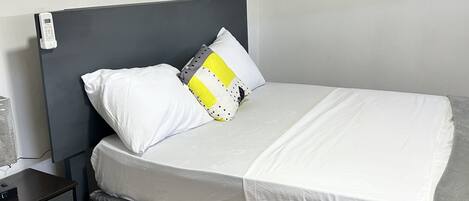 A neatly made bed with white bedding and a grey skirt, a bedside table with a phone, and a wooden floor.