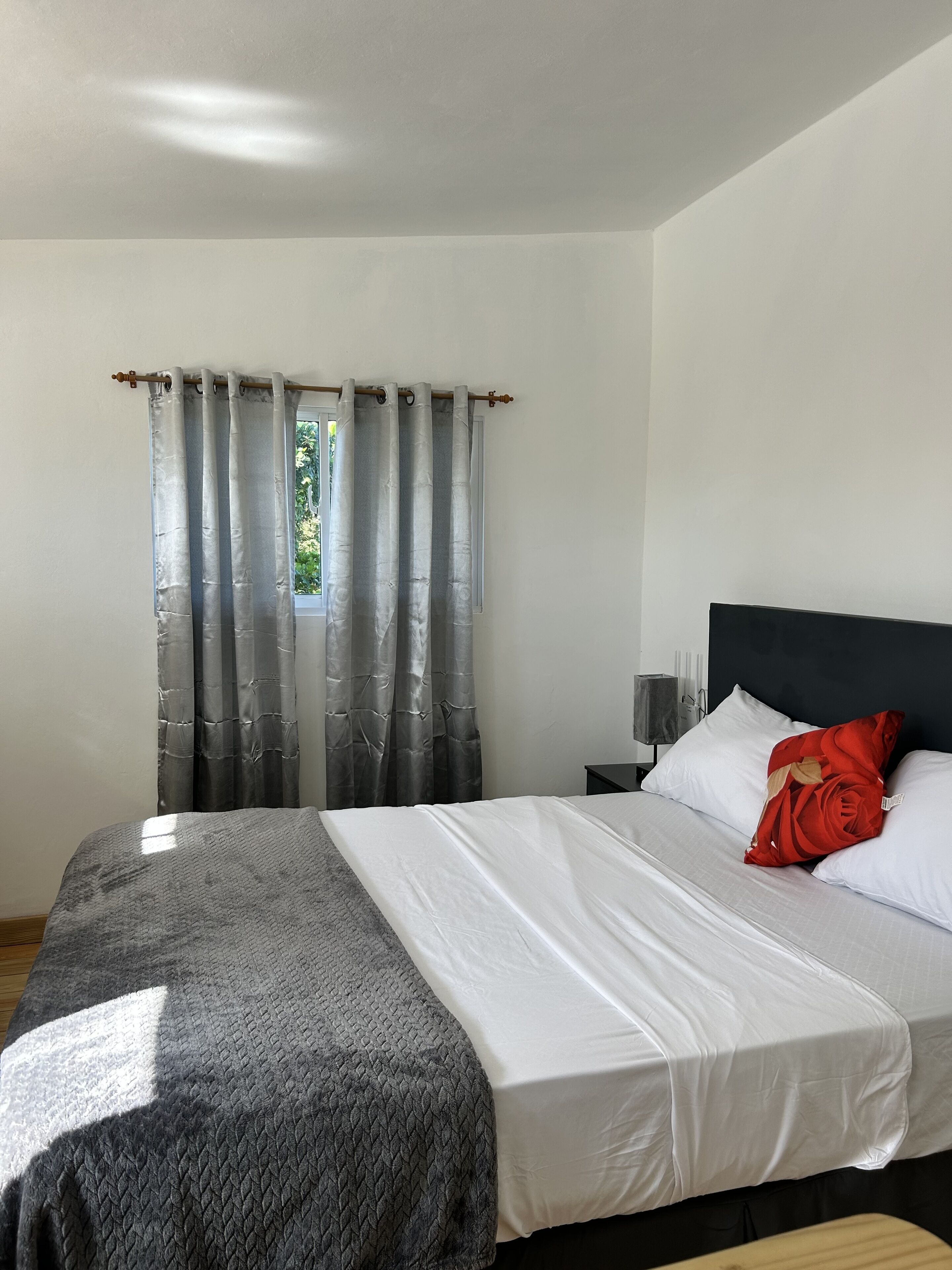 A bedroom with a bed, a nightstand, a window with curtains, and a view of greenery outside.