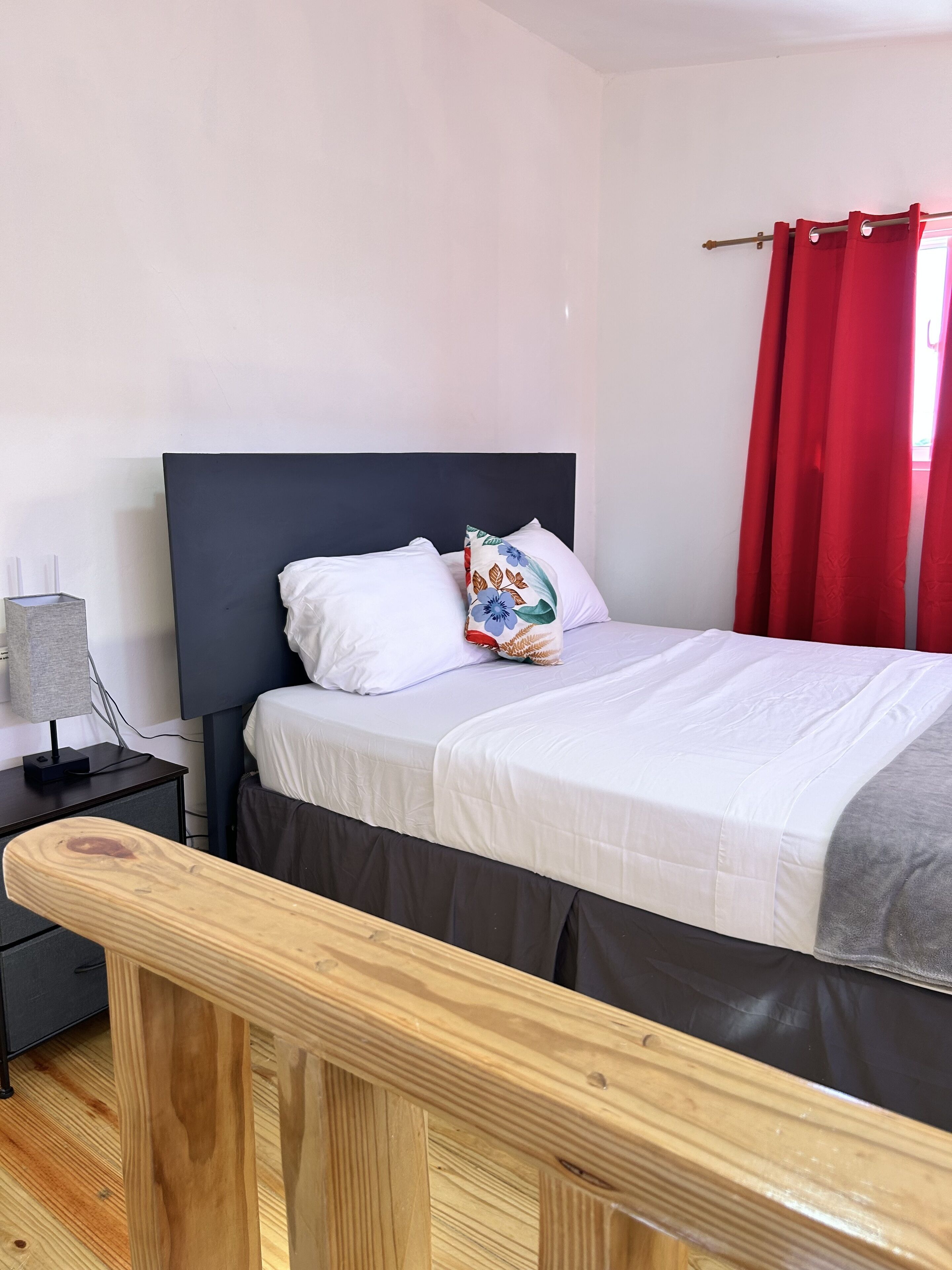 A bedroom with a bed, a nightstand, a lamp, and a window with red curtains.