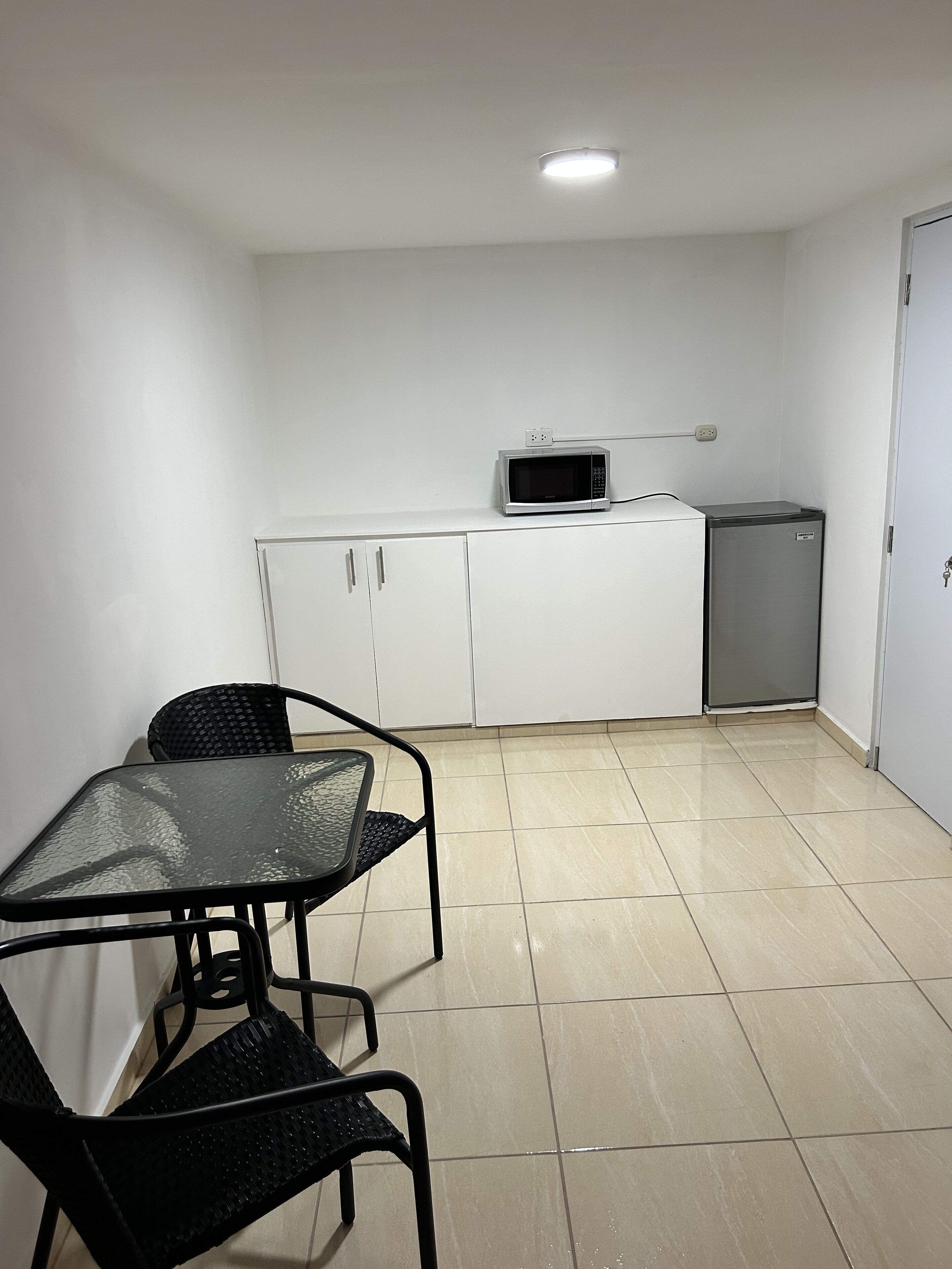 A small kitchenette with a microwave, a mini fridge, and white cabinets. There is a glass table with two chairs.