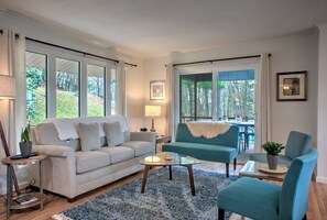 A living room with a sofa, armchairs, a coffee table, and a rug. There are windows with curtains, a lamp, and a framed picture on the wall.