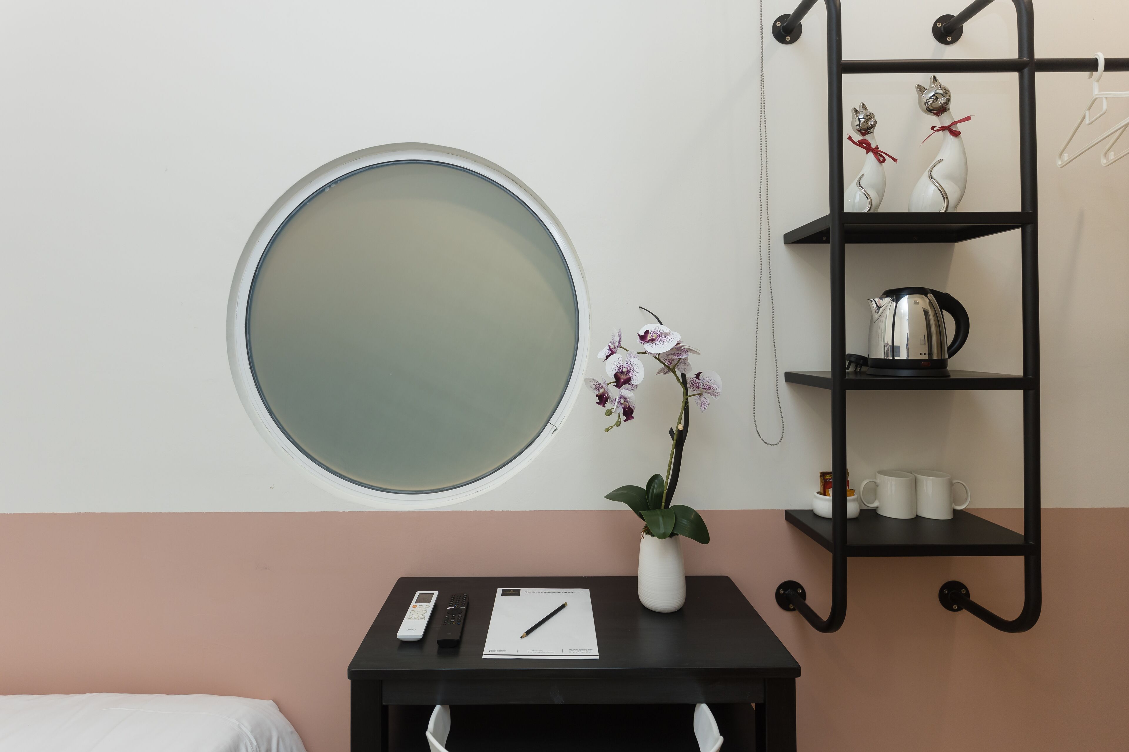 Une pièce avec un grand miroir rond, une petite table sur laquelle est posé un vase de fleurs, une étagère ornée d’objets décoratifs et une fenêtre.