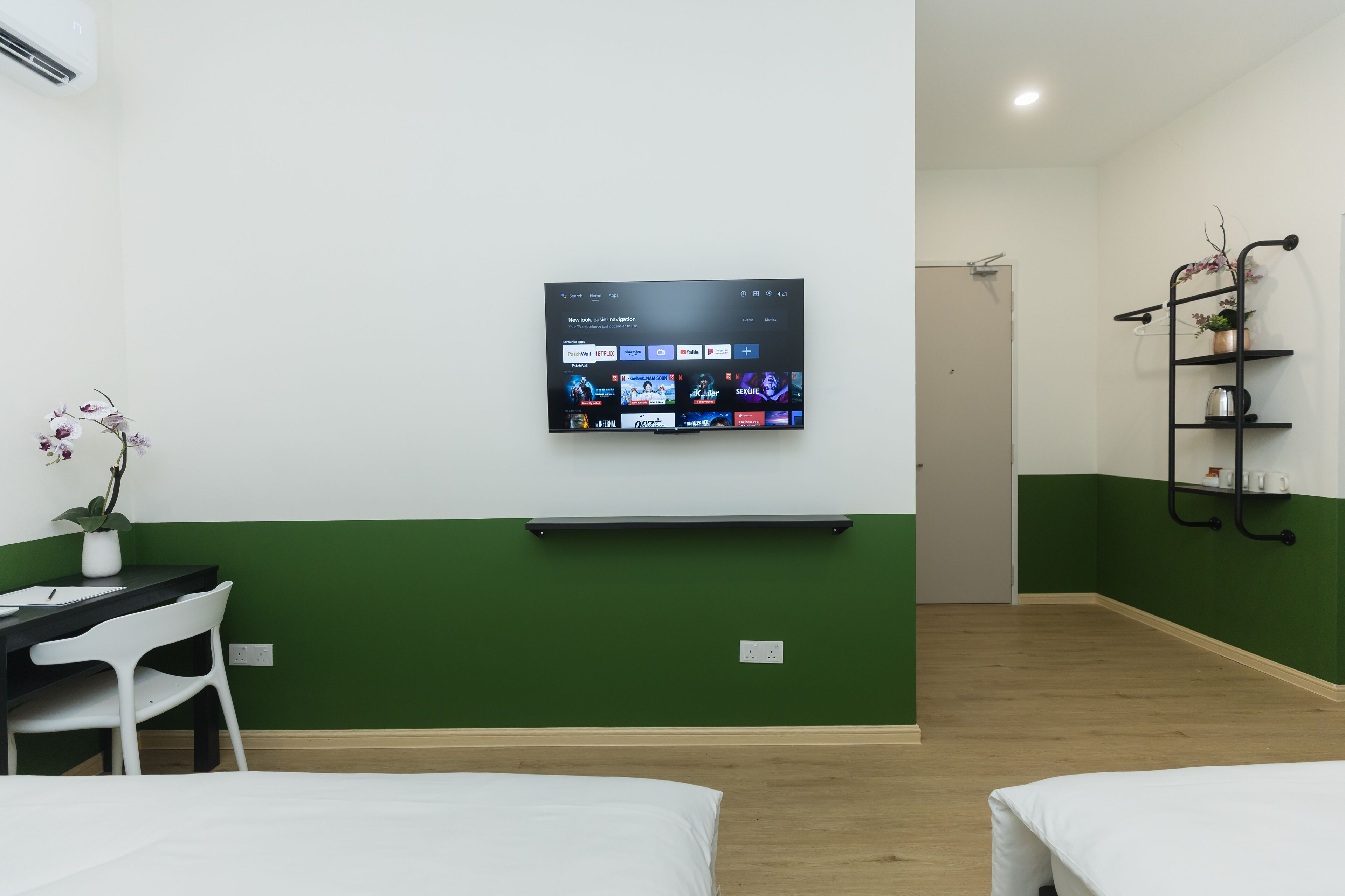 Habitación de hotel con televisión instalada en la pared mostrando diversas plataformas de streaming, un escritorio con silla, una planta en maceta y una cama con ropa blanca.