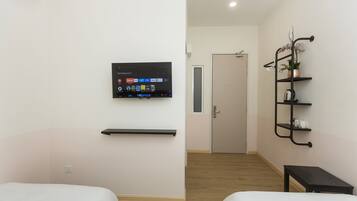 Habitación de hotel con dos camas, un televisor montado, un estante con una planta en maceta y una puerta que da a otra habitación.