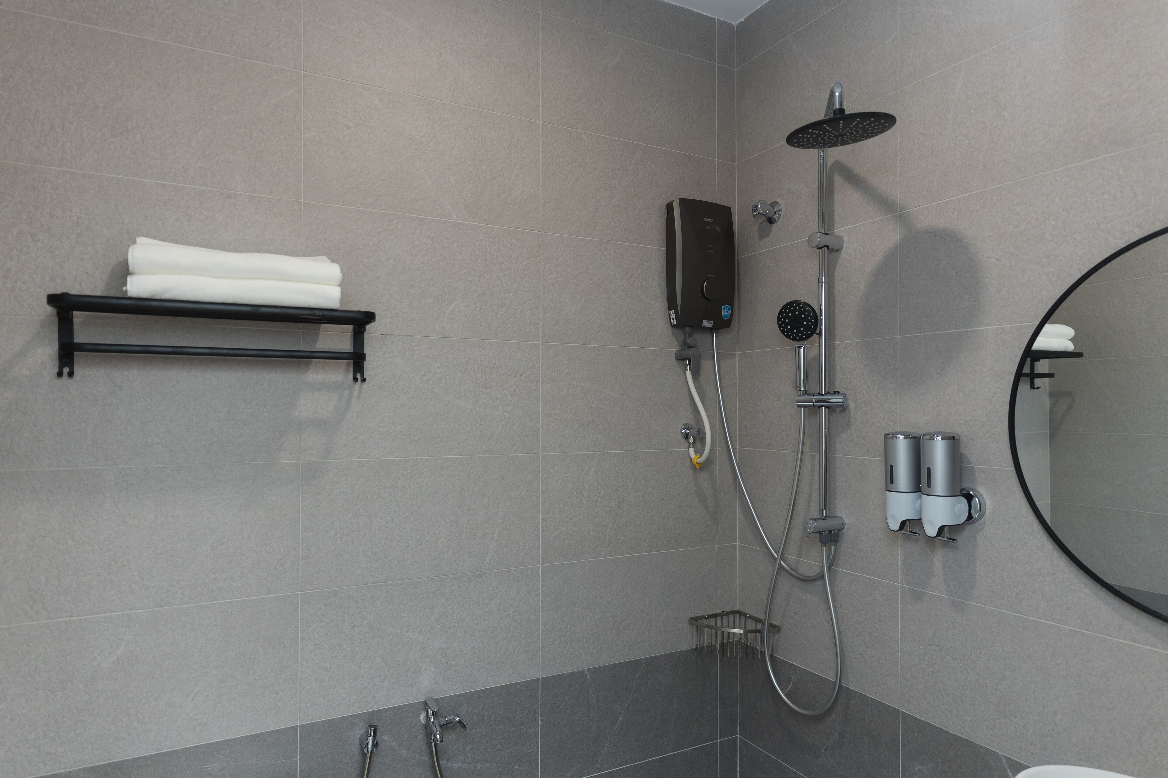 A modern bathroom with grey tiles, a black towel rack, a handheld showerhead, a wall-mounted heater, and a round mirror.
