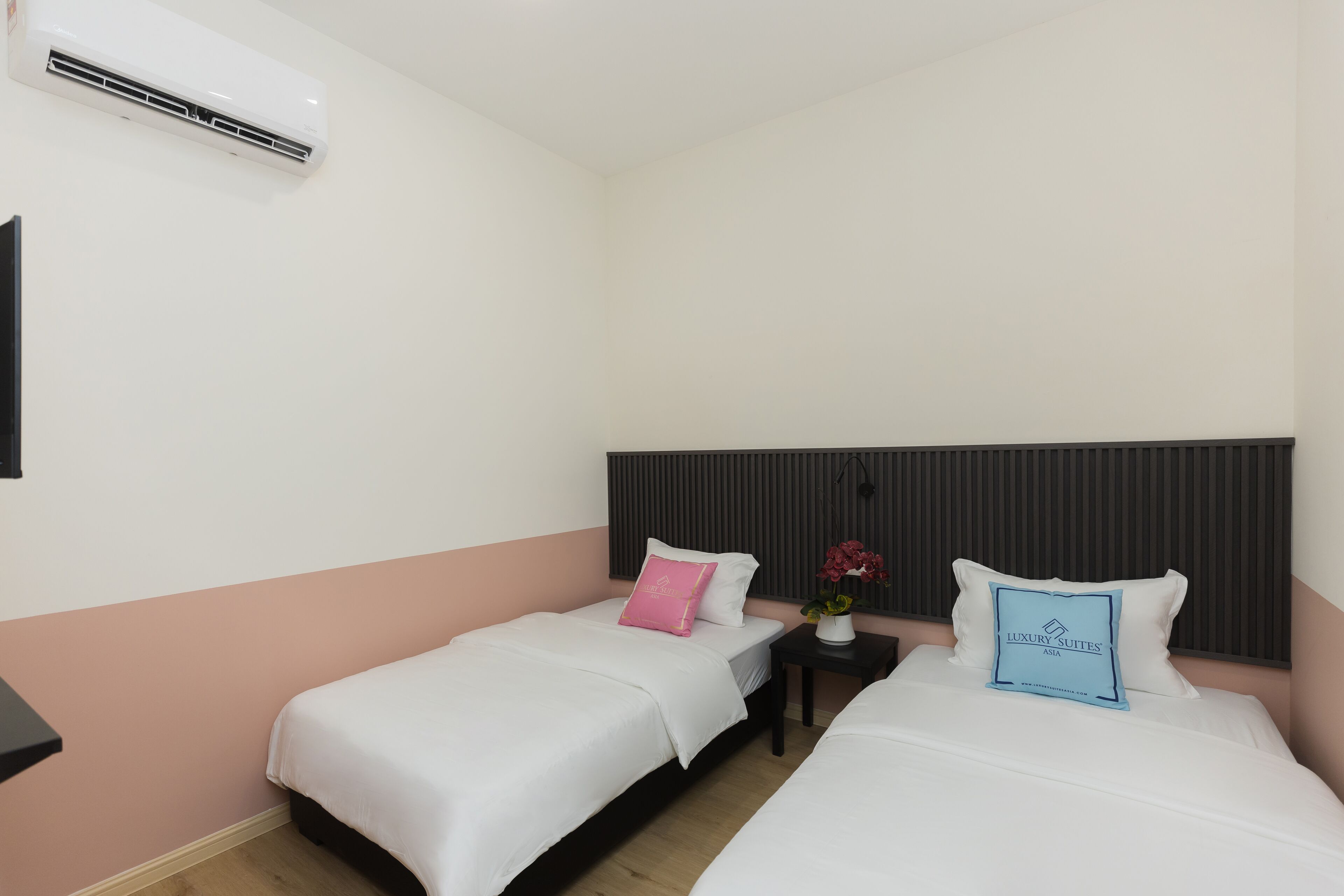 A hotel room with two beds, a bedside table with a vase of flowers, and a wall-mounted air conditioner.