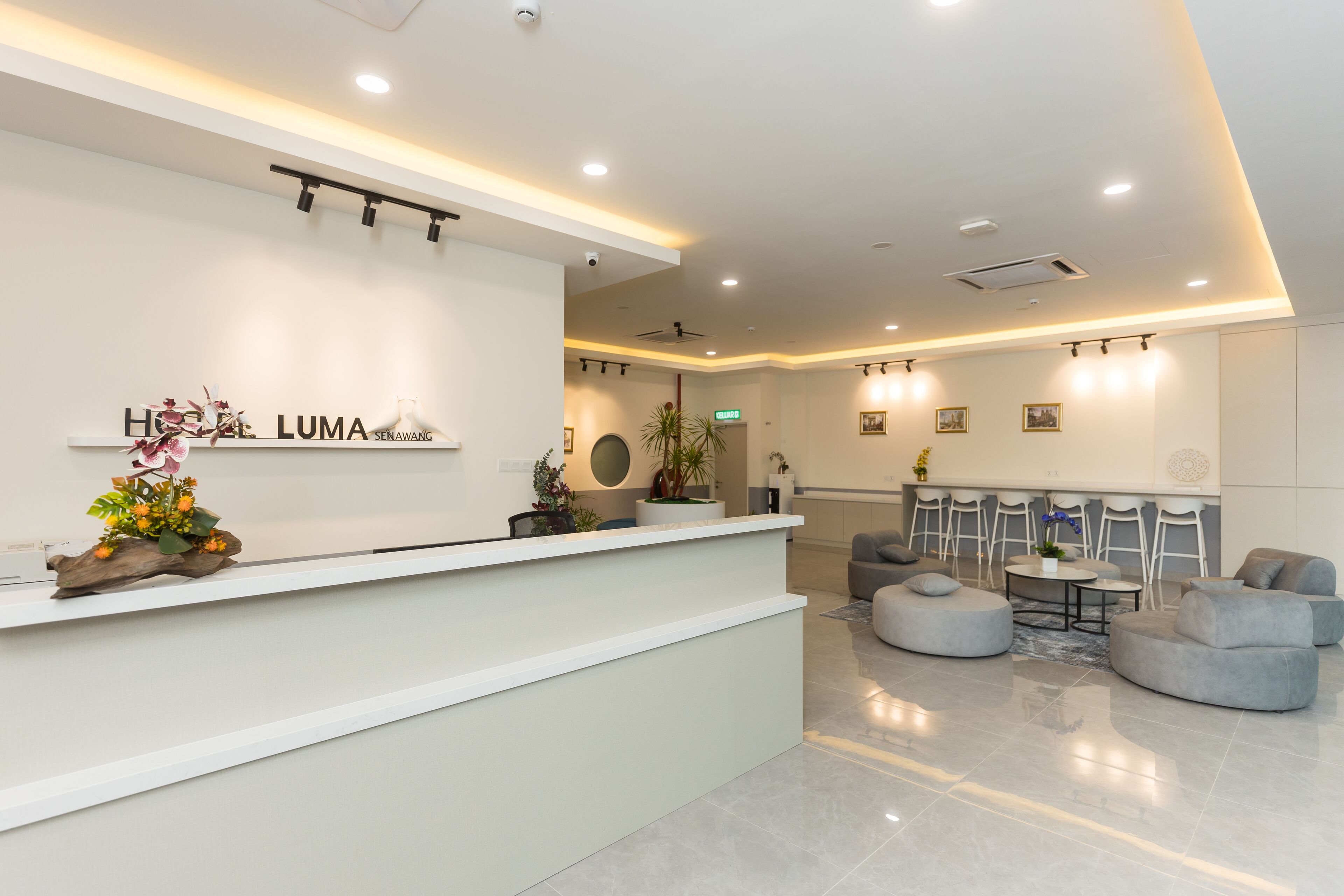 A modern hotel reception area with a white reception desk, a seating area with grey sofas, and a dining table.