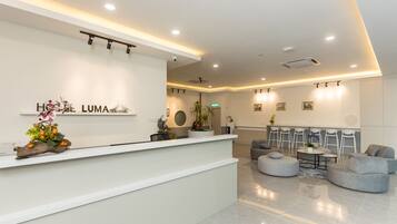 A modern hotel reception area with a white reception desk, a seating area with grey sofas, and a dining table.