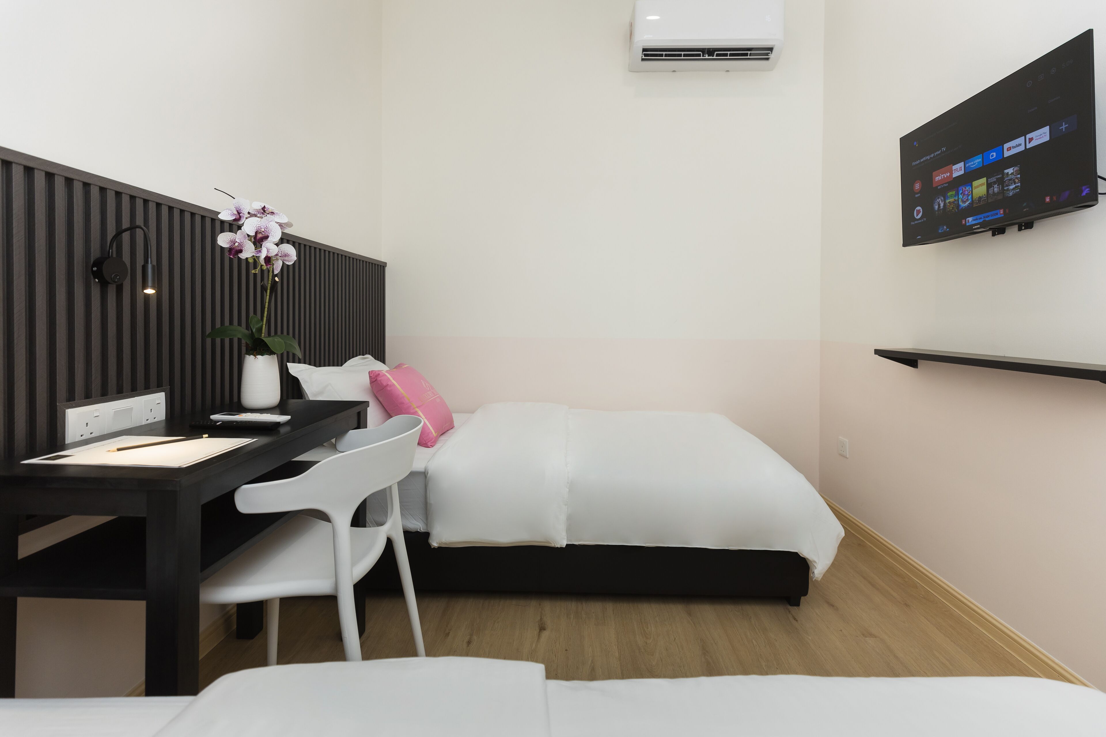 Una habitación de hotel moderna con un escritorio de madera, una silla blanca, una cama con ropa de cama blanca, una pequeña orquídea en maceta, un televisor de pantalla plana montado en la pared y un equipo de aire acondicionado.