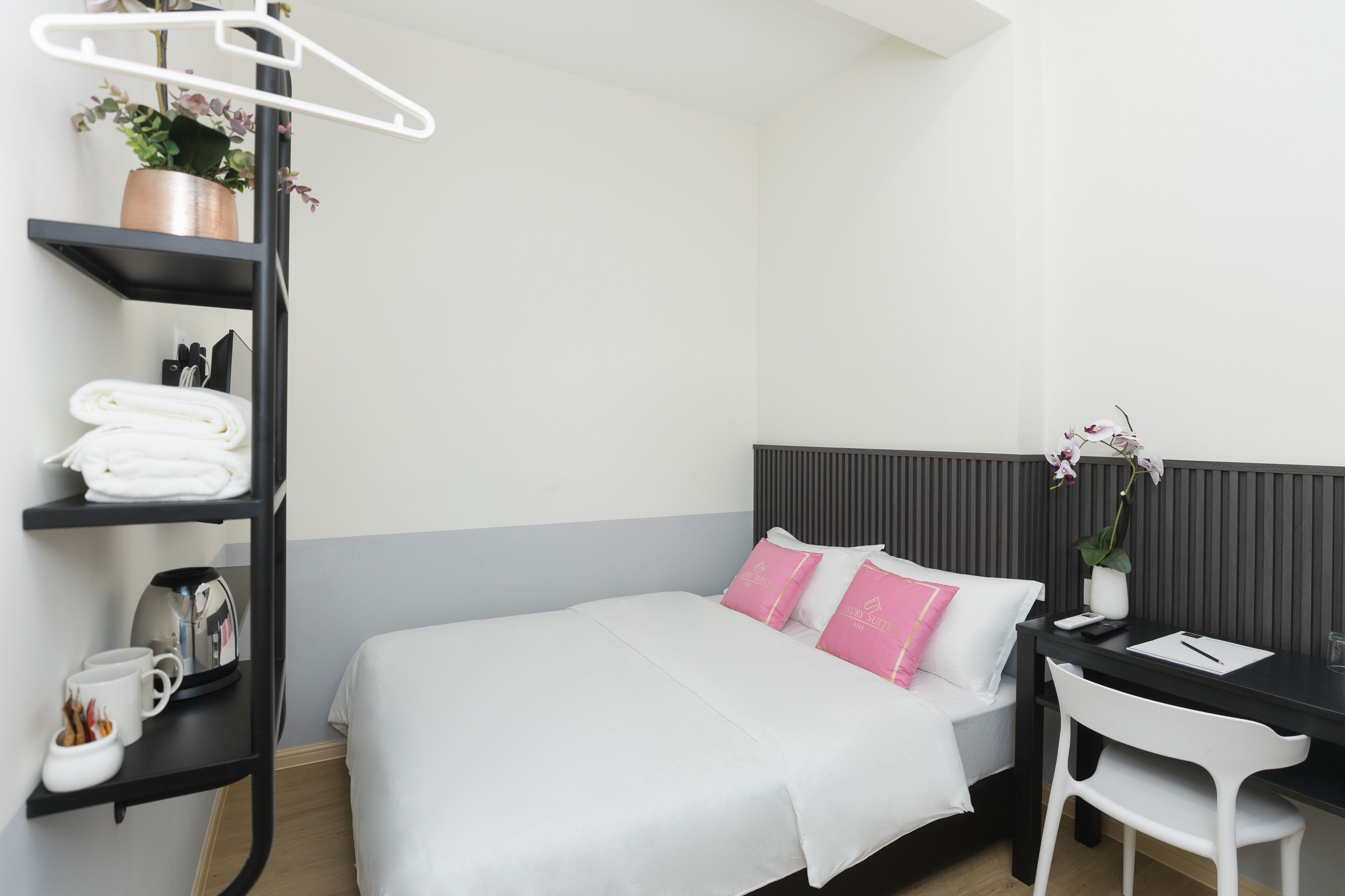 A neatly arranged hotel room with a bed, a desk, and a chair. There are shelves with towels and a coffee maker.