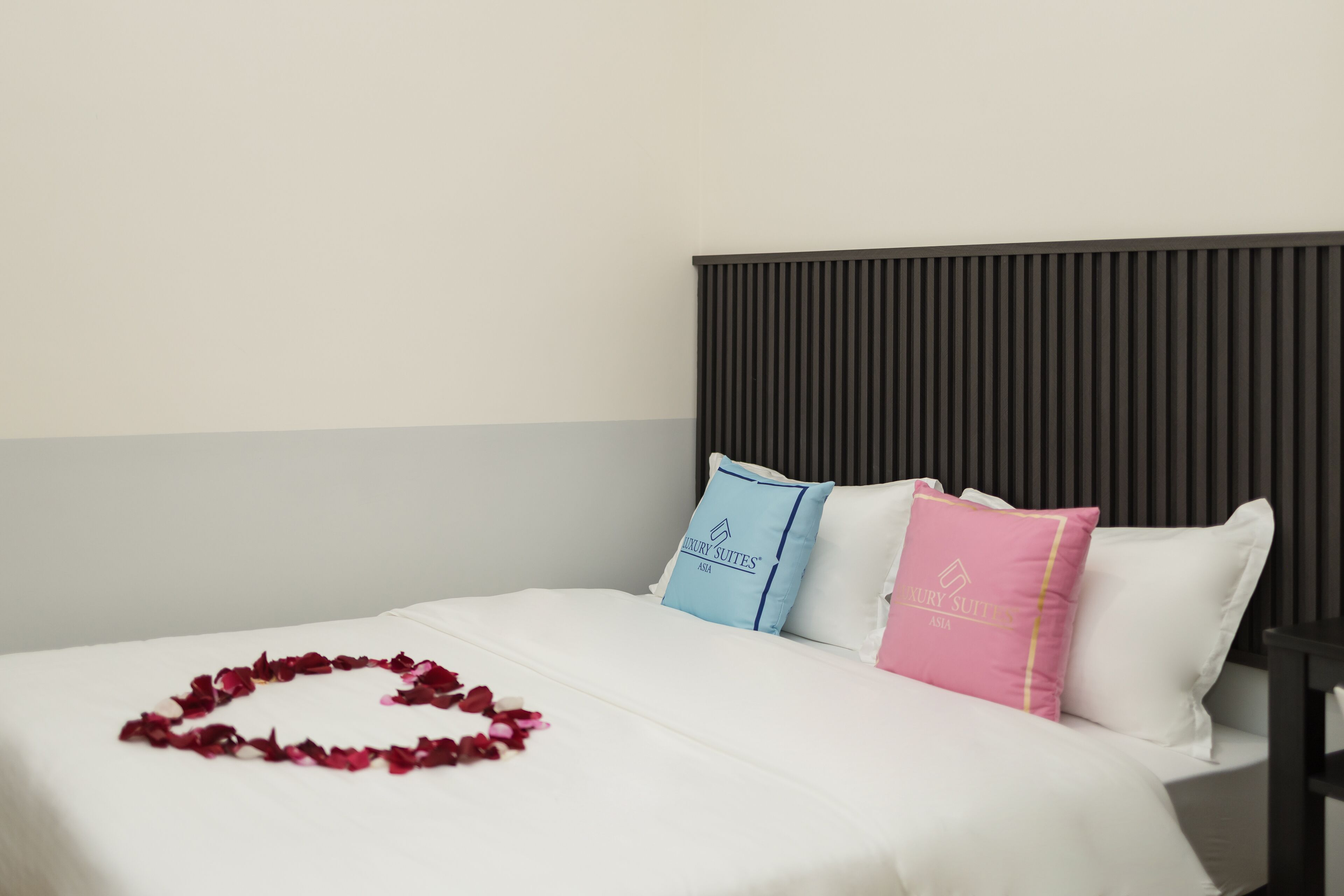 A neatly made bed with a heart-shaped arrangement of rose petals, two pillows with branding, and a dark headboard.