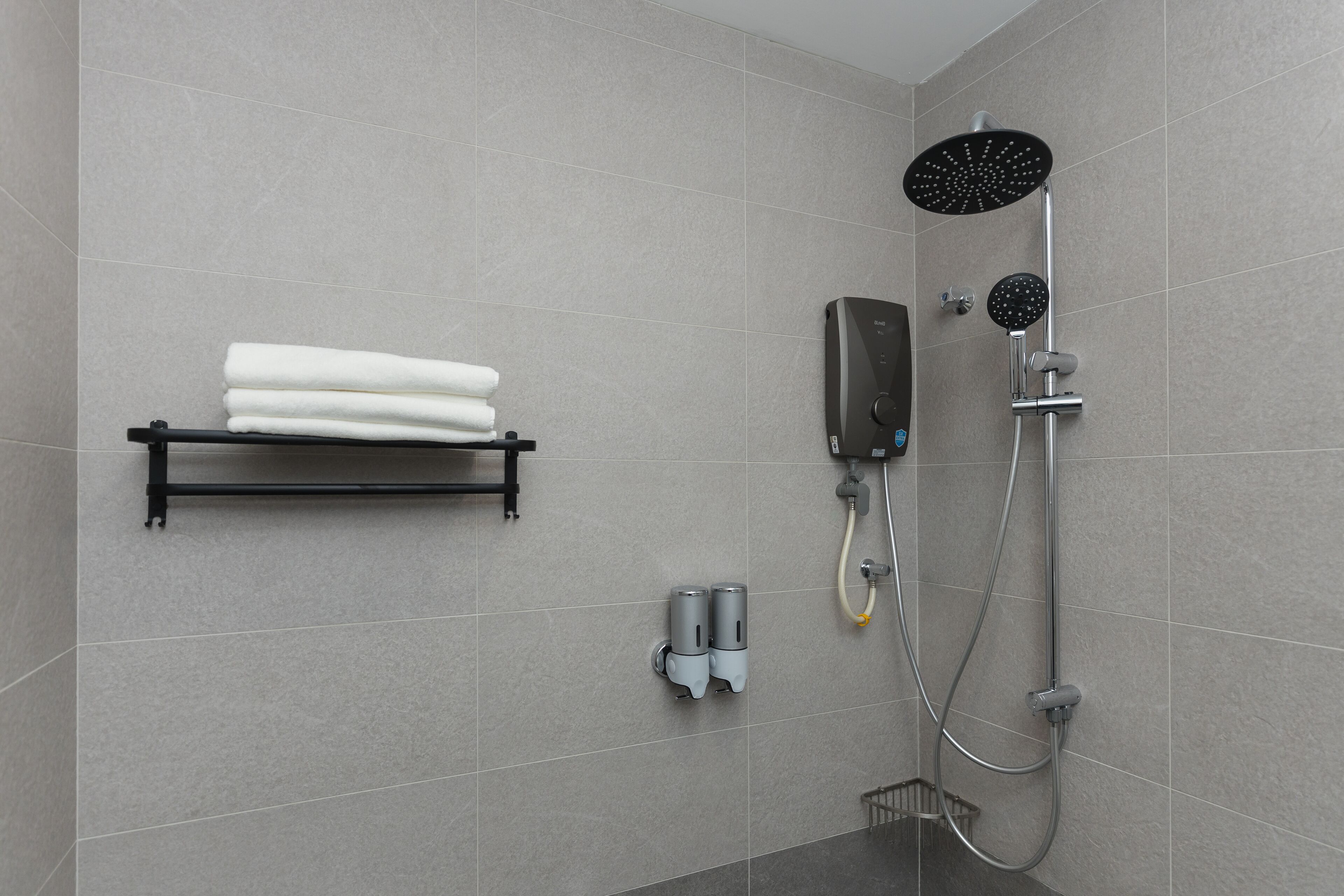 A modern bathroom with a grey tiled wall, a black towel rack with folded towels, a handheld showerhead, and a wall-mounted soap dispenser.