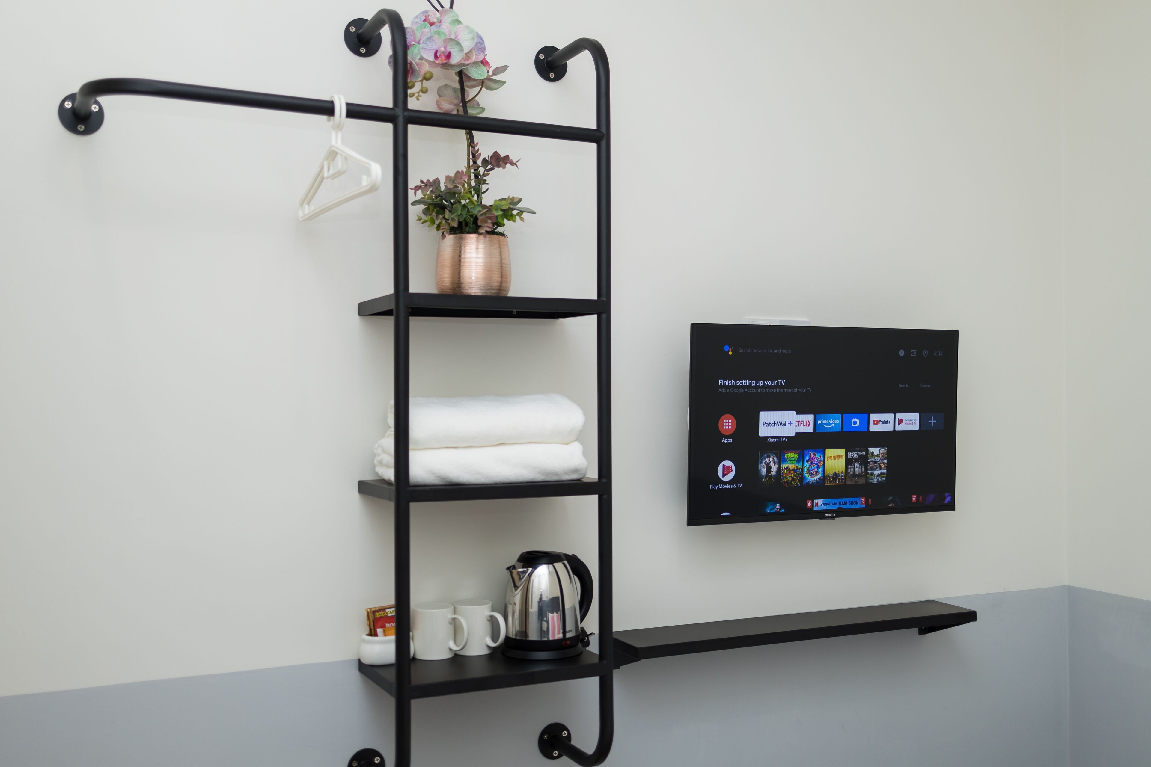 A black metal shelf unit with a flat-screen TV mounted on the wall, holding a plant, folded towels, and a kettle.
