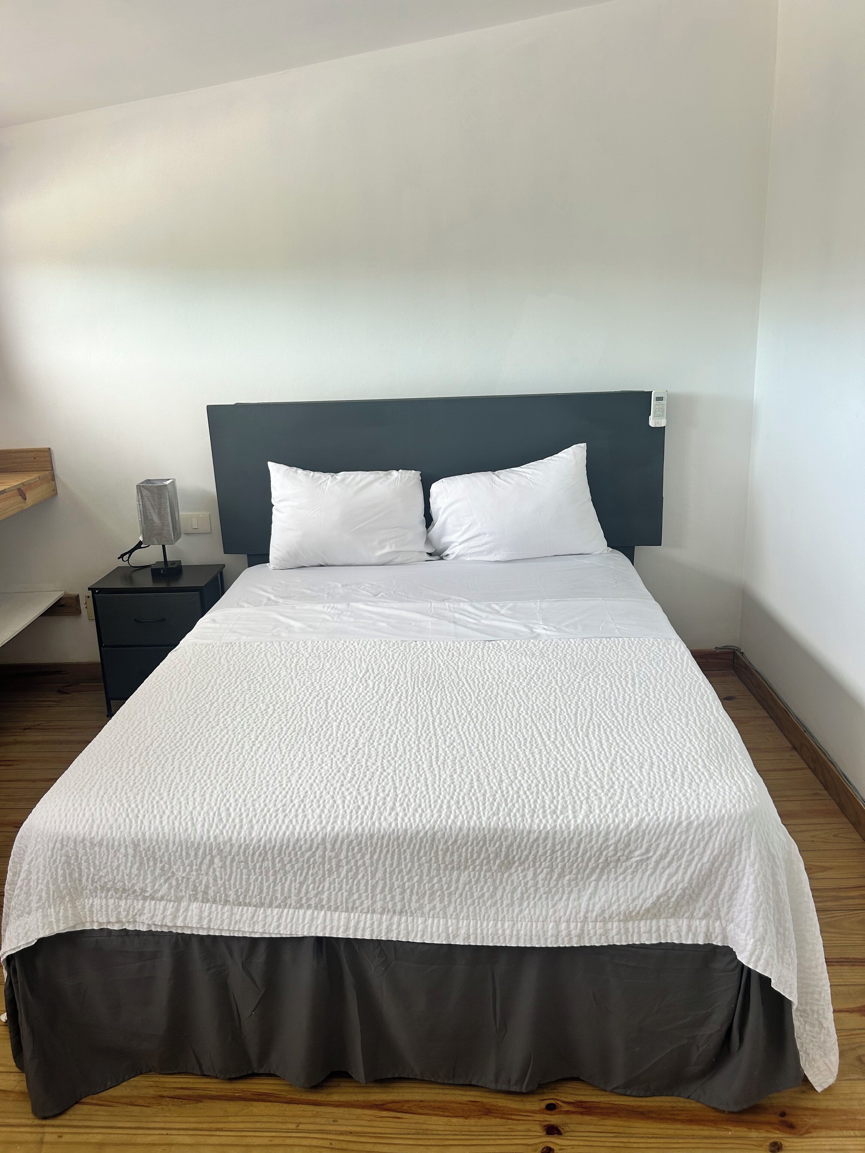 Una cama bien hecha con ropa de cama blanca y un cabecero oscuro, una mesita de noche con lámpara y suelo de madera.