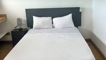 Una cama bien hecha con ropa de cama blanca y un cabecero oscuro, una mesita de noche con lámpara y suelo de madera.