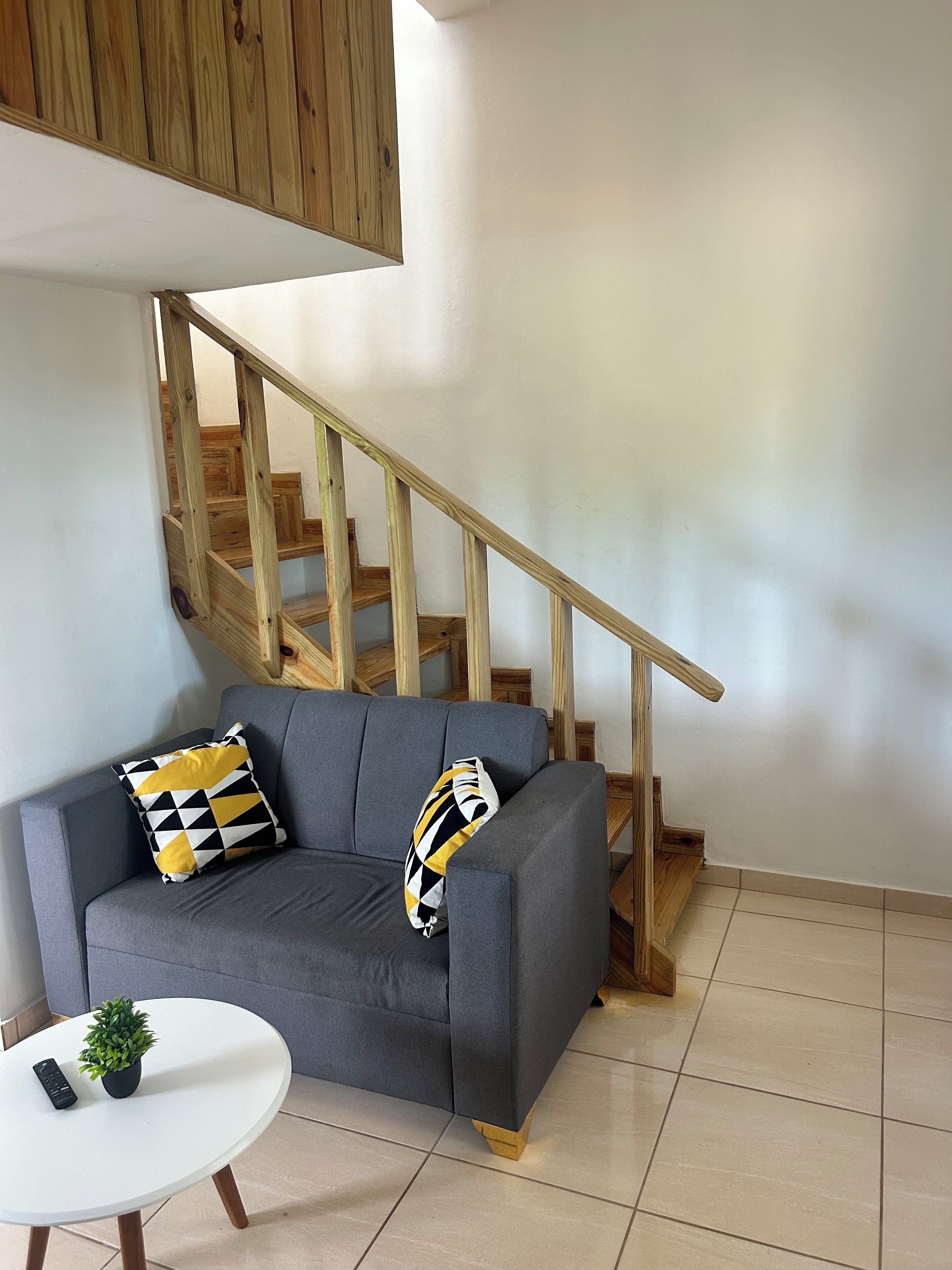 A grey sofa with two patterned cushions, a small white table with a potted plant, and a wooden staircase.