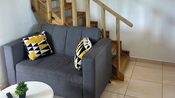 Un sofá gris con dos cojines estampados, una mesita blanca con una planta en maceta, y una escalera de madera.