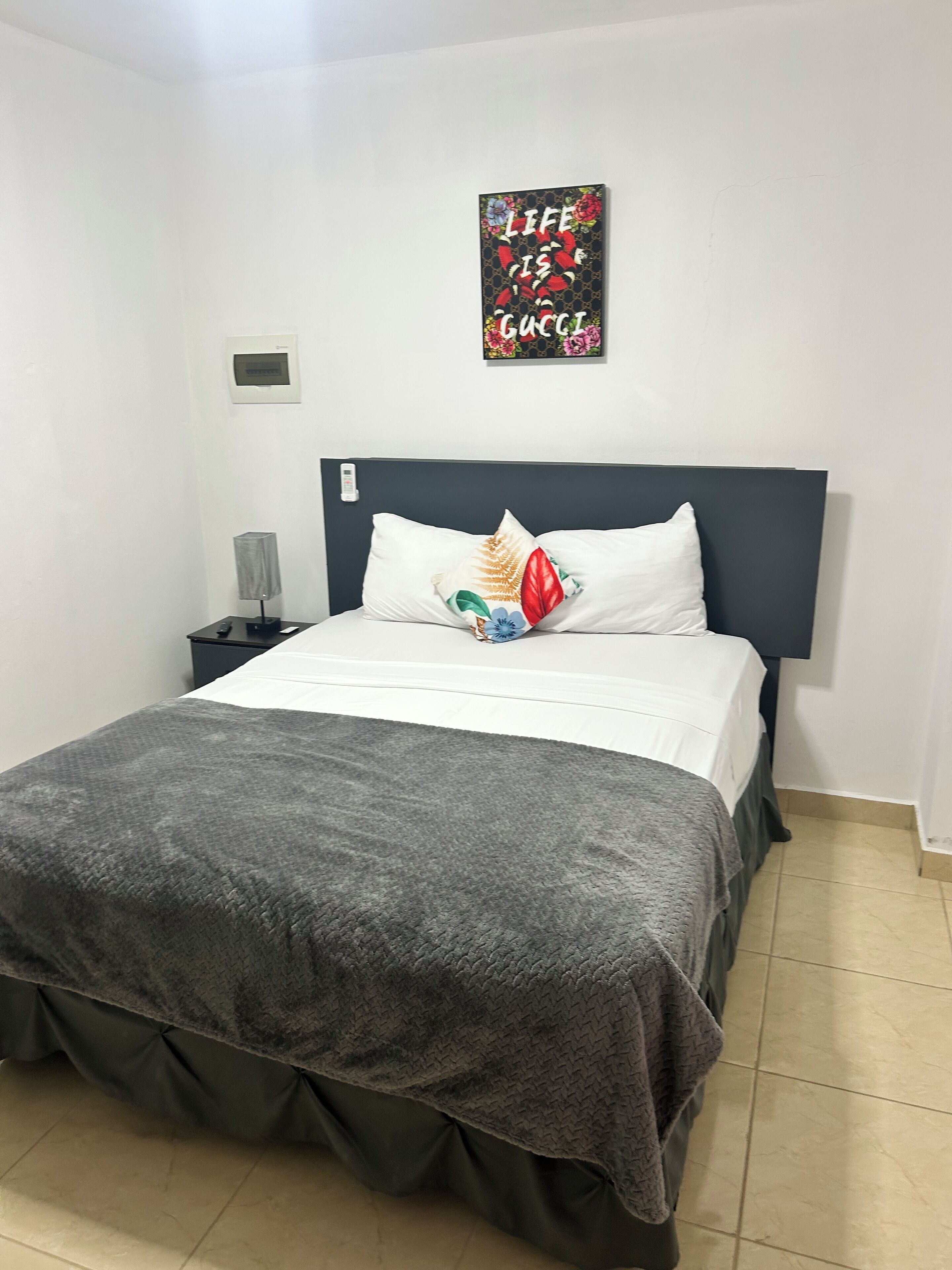 Una cama bien hecha con un edredón gris, almohadas blancas y una almohada decorativa con motivos florales. Se ve una mesita de noche con una lámpara y un termostato en la pared.