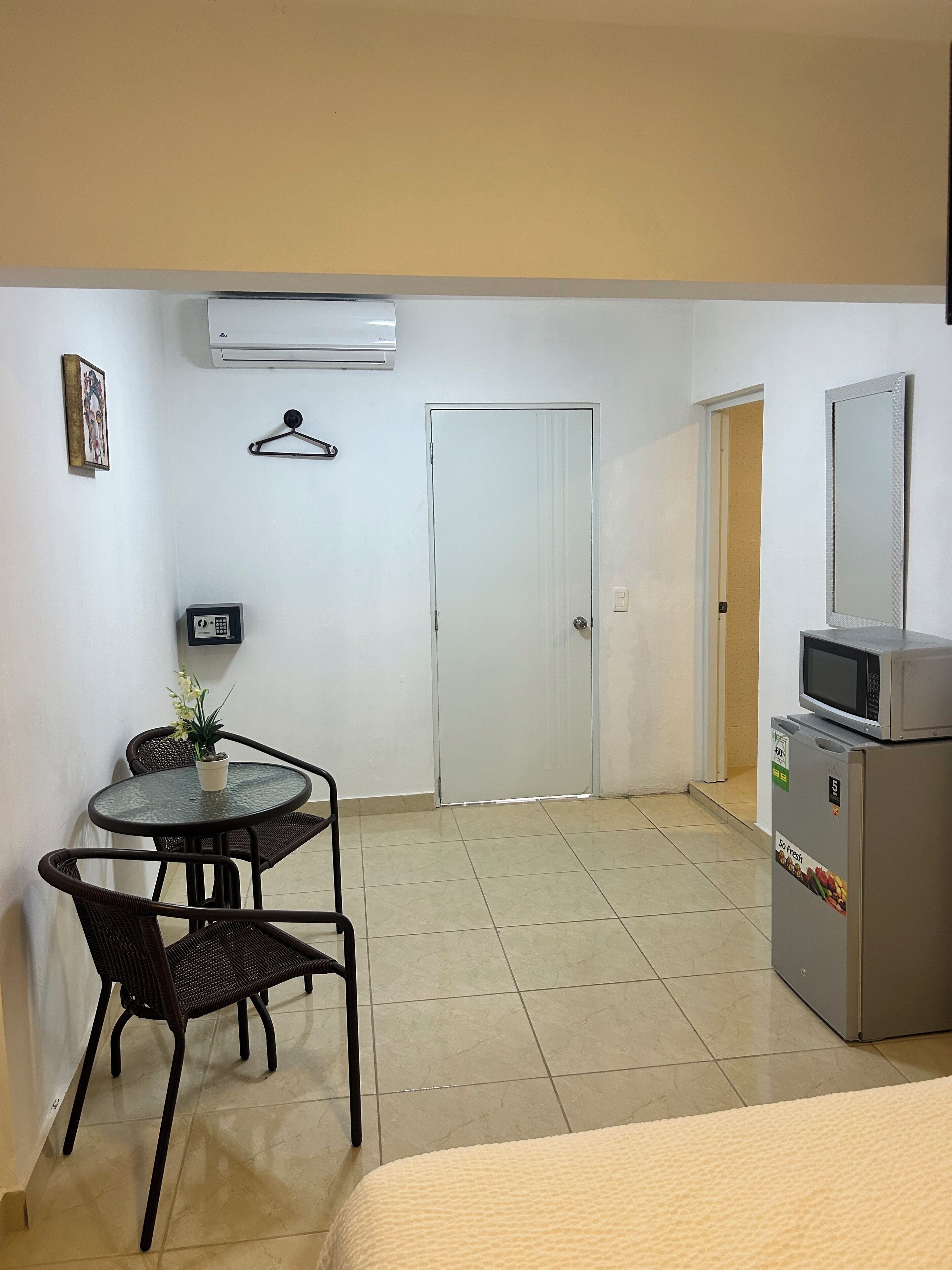 A hotel room with a bed, a small round table with chairs, a potted plant, an air conditioner, a door, a refrigerator, and a wall-mounted TV.
