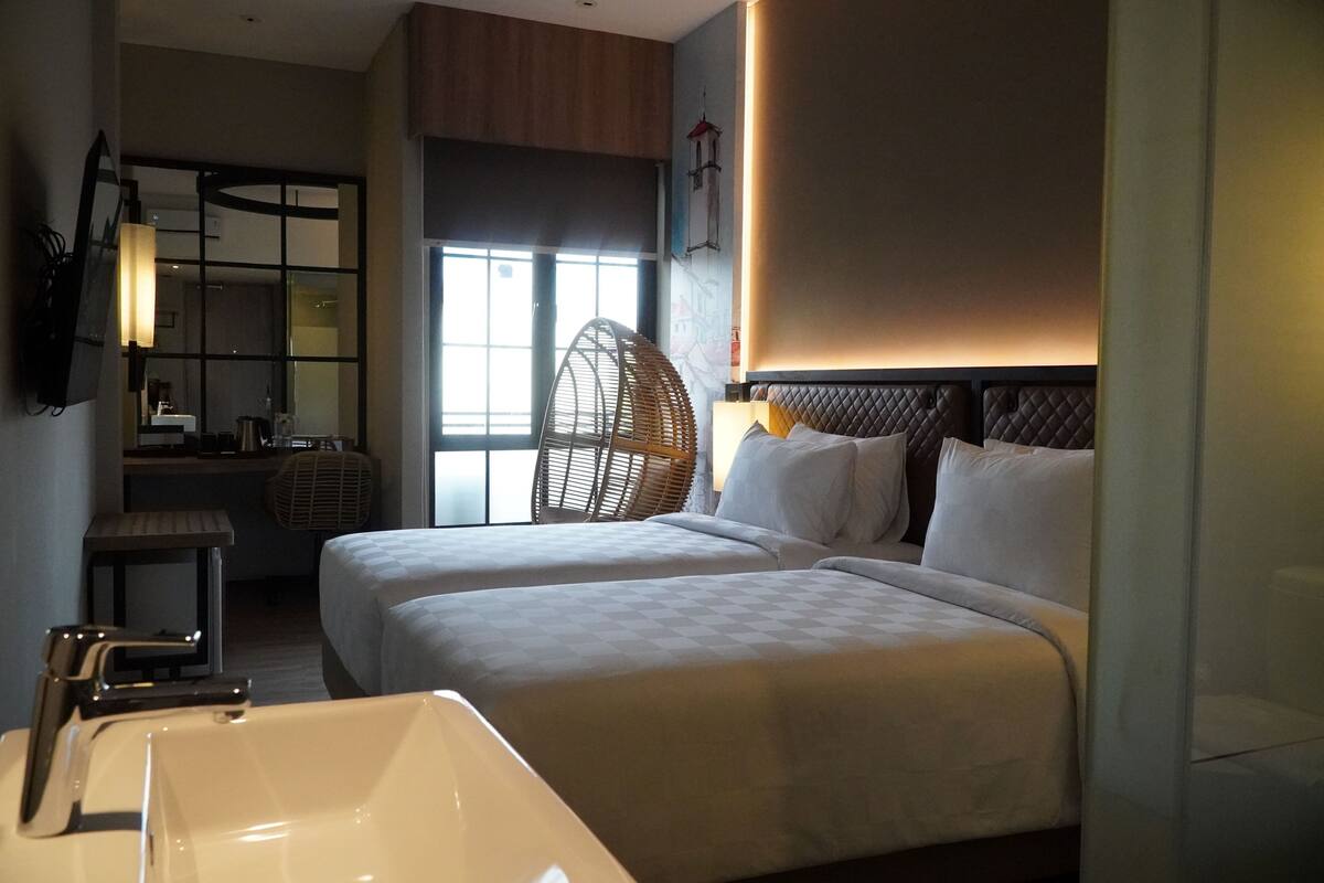 Une chambre d’hôtel avec deux lits, une grande fenêtre, un coin salon avec un fauteuil, une petite table et une salle de bain avec un lavabo et un miroir.