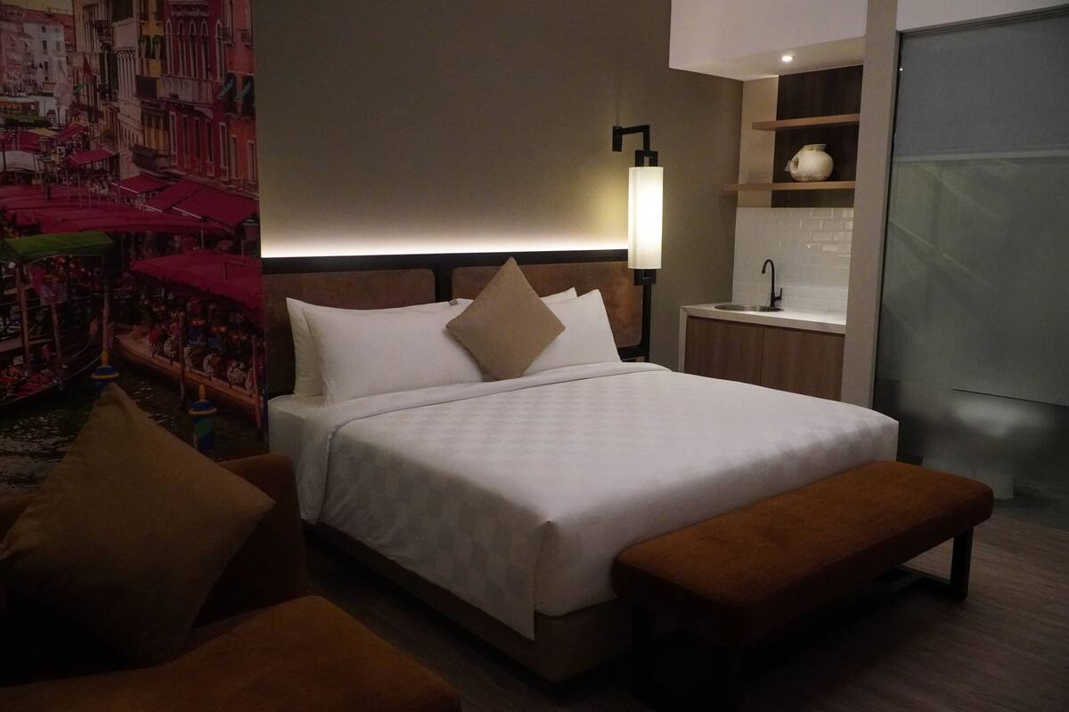 Une chambre d’hôtel comprenant un lit, un banc, une table de chevet, une lampe fixée au mur, un lavabo et une étagère avec un vase.