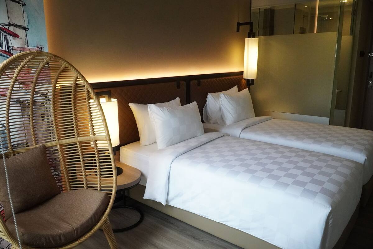 Une chambre d’hôtel avec un lit, deux oreillers, une table de chevet, une lampe et une chaise en osier.