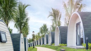 Una hilera de cabañas modernas con techo blanco y revestimiento oscuro, alineadas a lo largo de un camino pavimentado, con palmeras al fondo.