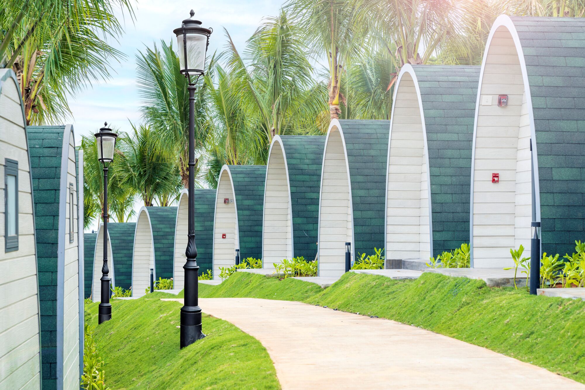 Un sendero conduce a una hilera de edificios modernos con techos blancos y revestimiento verde, rodeados de abundante vegetación y palmeras.