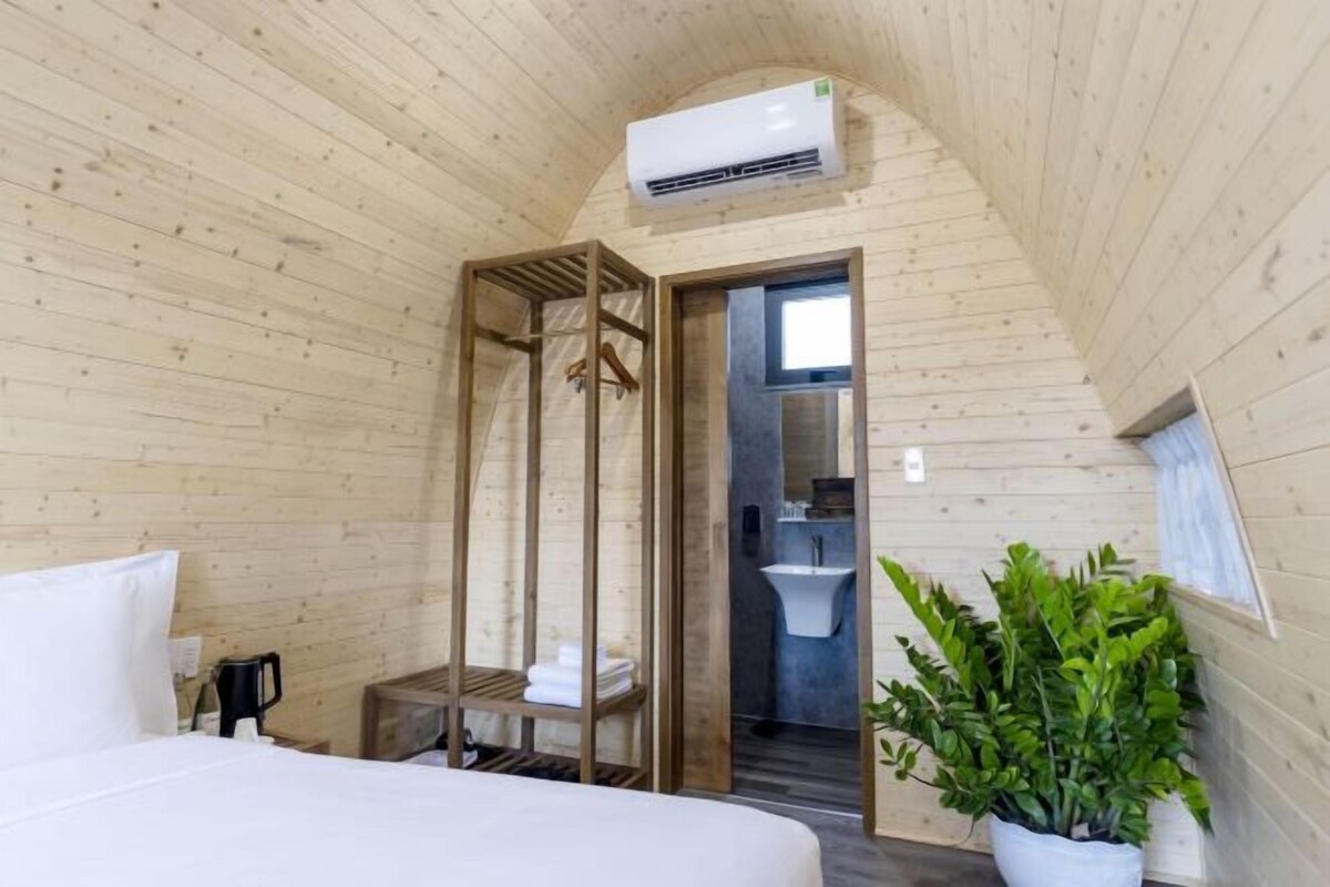 Una habitación con cama, un armario de madera, una planta en maceta y un baño visible a través de una puerta abierta.