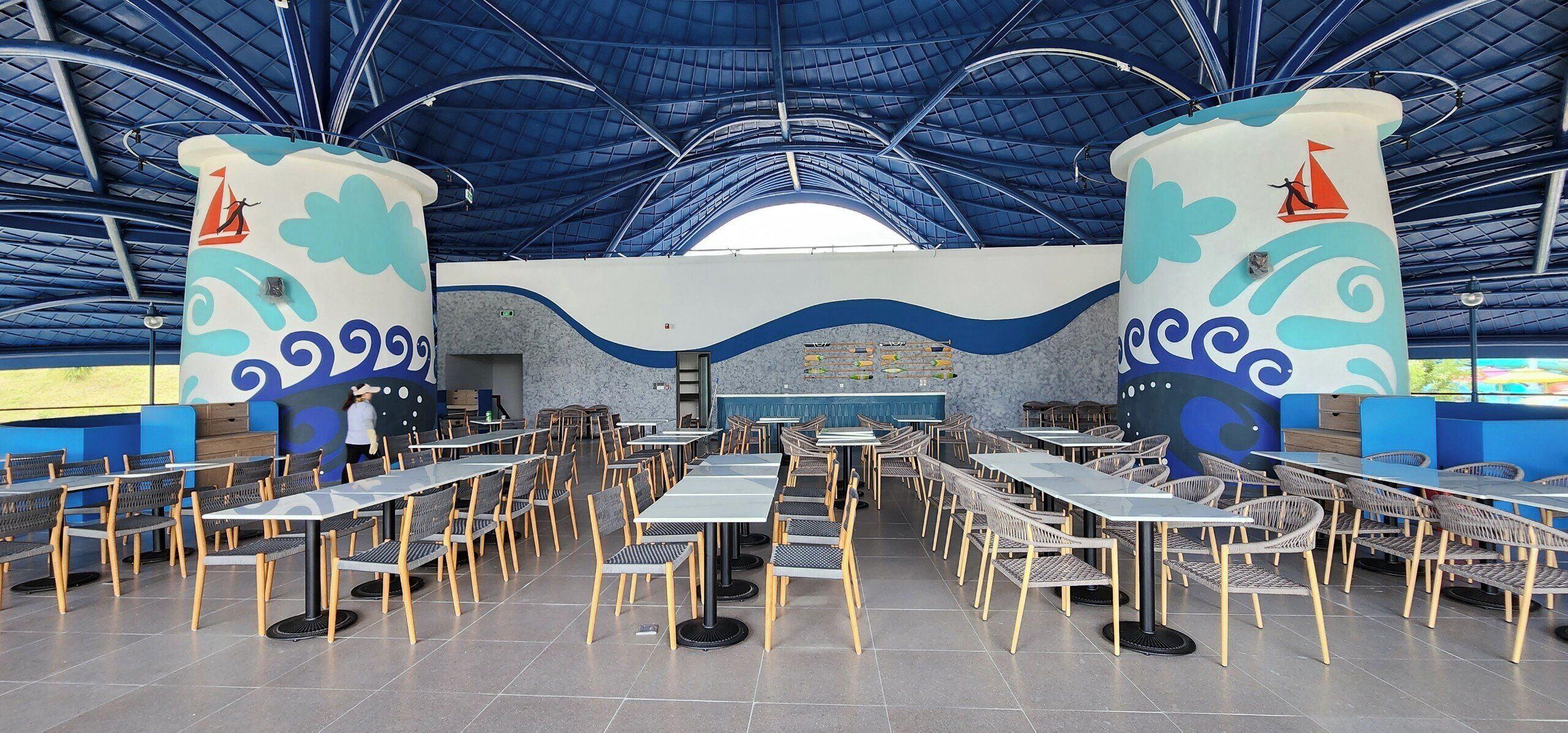 Un comedor interior espacioso con techos altos y decoración temática náutica. Mesas y sillas están dispuestas en filas, y hay dos pilares grandes con diseños de veleros.