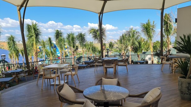 Un patio al aire libre con mesas redondas y sillas de mimbre, con vistas a una piscina y palmeras.
