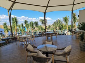Une terrasse extérieure avec des tables rondes et des chaises en osier, donnant sur une piscine et des palmiers.