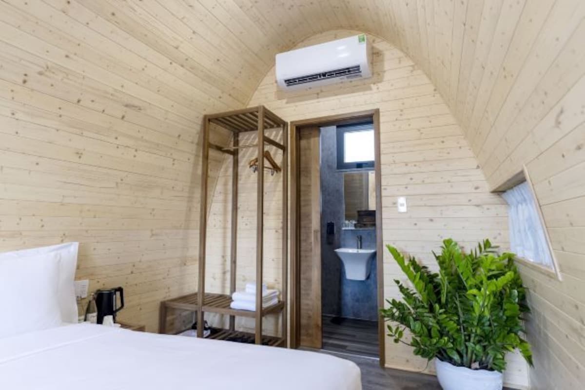 Un dormitorio de cabaña de madera con cama, una mesita, un perchero, una planta en maceta y un baño con lavamanos y espejo.