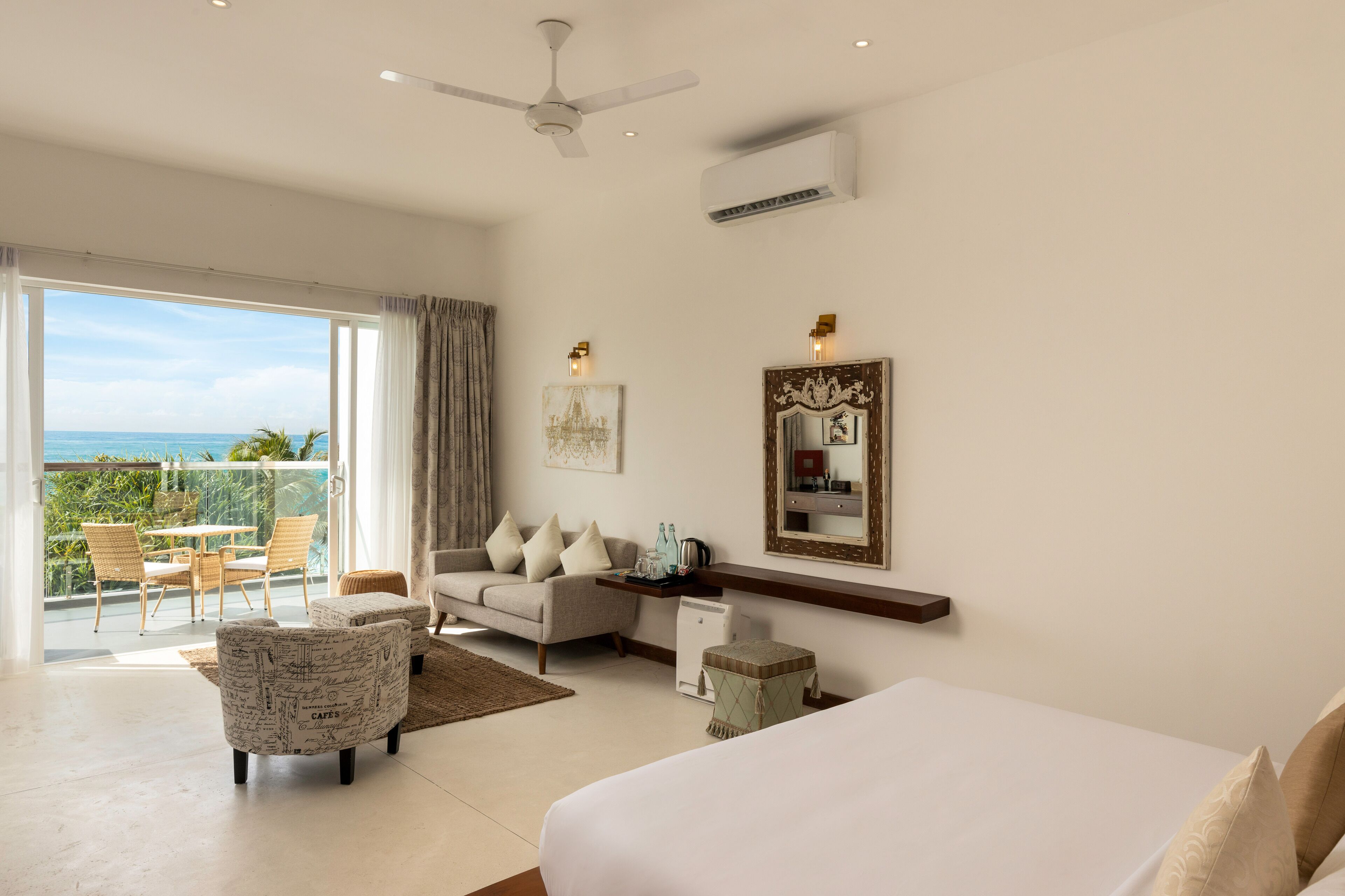 A spacious bedroom with a large bed, a sofa, a dining area with chairs, and a balcony with a view of the ocean.