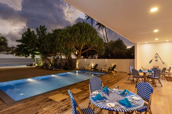 Un área de piscina al aire libre con sillones de descanso y mesas, vista al océano y una terraza de madera.
