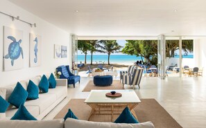 A modern living room with a view of the beach, featuring a white sofa, blue armchairs, and a glass coffee table. - Vis Ta Vie -Thema Collection (Kamburugamuwa)